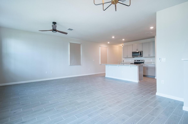 kitchen with gray cabinets, appliances with stainless steel finishes, ceiling fan with notable chandelier, and an island with sink