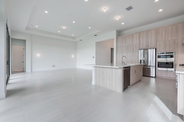 kitchen with a center island with sink, light tile patterned floors, a tray ceiling, and appliances with stainless steel finishes