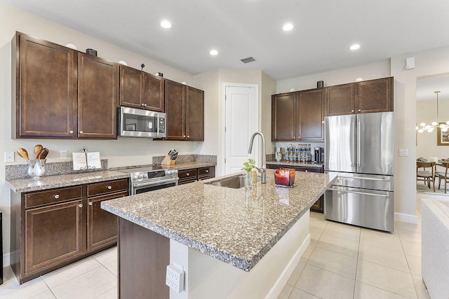 kitchen with appliances with stainless steel finishes, sink, a kitchen island with sink, light tile patterned flooring, and light stone counters