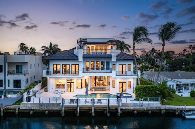 back house at dusk with a balcony, a pool with hot tub, a water view, and a patio