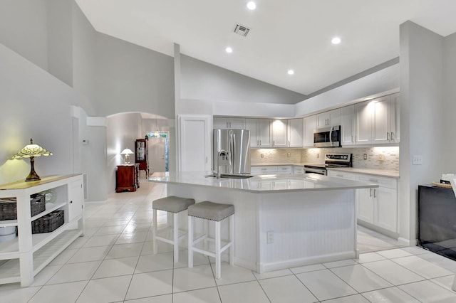 kitchen featuring light tile patterned floors, appliances with stainless steel finishes, white cabinetry, high vaulted ceiling, and a center island with sink