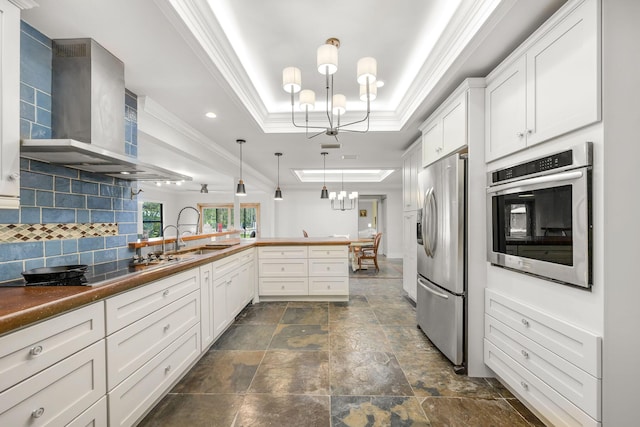 kitchen with wall chimney range hood, hanging light fixtures, stainless steel appliances, kitchen peninsula, and a raised ceiling