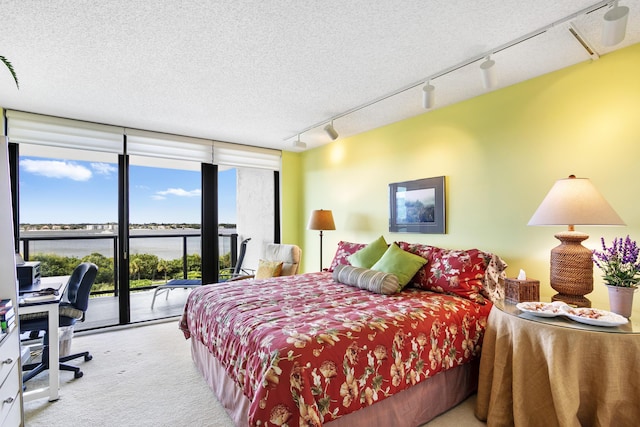 carpeted bedroom with rail lighting, a water view, a textured ceiling, access to outside, and a wall of windows