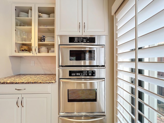 kitchen with white cabinetry, stainless steel double oven, tasteful backsplash, and dark stone countertops