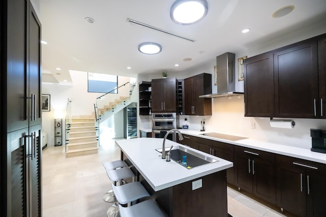 kitchen featuring black electric stovetop, a center island with sink, sink, a kitchen breakfast bar, and wall chimney exhaust hood