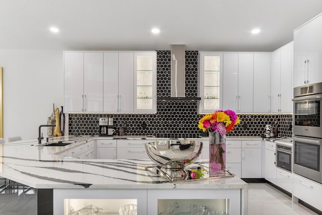 kitchen with wall chimney exhaust hood, white cabinets, light stone counters, and stainless steel double oven