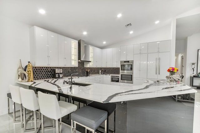 kitchen with lofted ceiling, kitchen peninsula, wall chimney exhaust hood, and white cabinetry