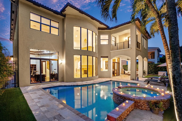 back house at dusk with a balcony, a patio area, and a pool with hot tub