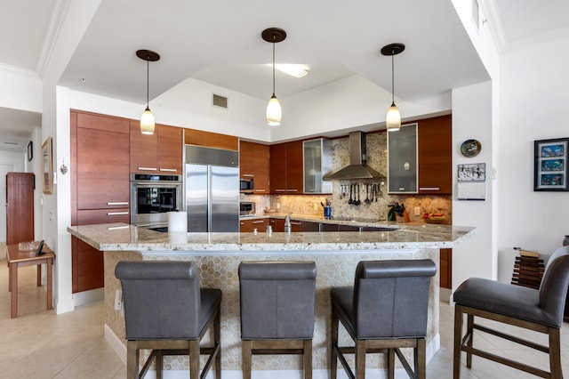 kitchen featuring appliances with stainless steel finishes, a breakfast bar area, backsplash, hanging light fixtures, and wall chimney range hood