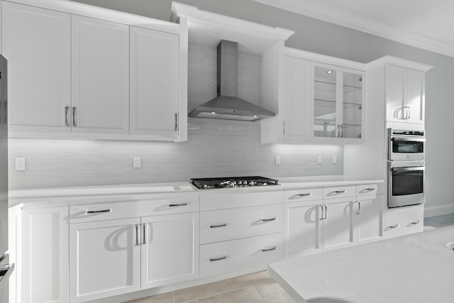 kitchen with appliances with stainless steel finishes, tasteful backsplash, white cabinetry, ornamental molding, and wall chimney range hood