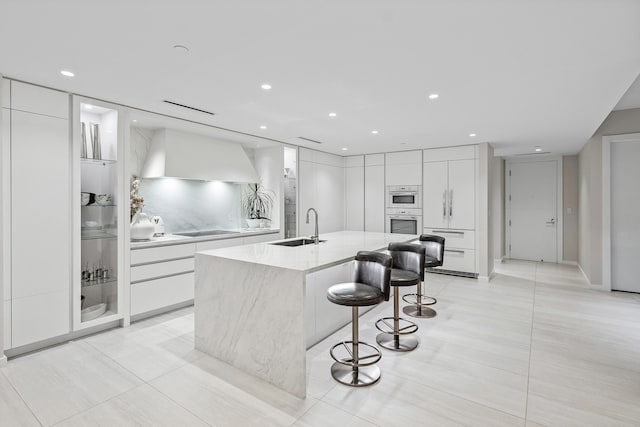 kitchen featuring white cabinets, wall chimney range hood, a center island with sink, and sink