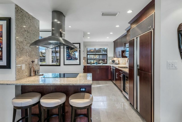 kitchen with light stone counters, island range hood, kitchen peninsula, black electric stovetop, and backsplash