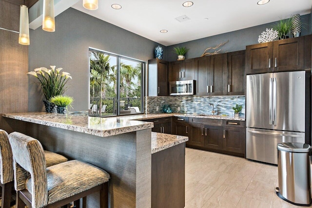 kitchen with dark brown cabinets, stainless steel appliances, light stone countertops, a kitchen bar, and kitchen peninsula