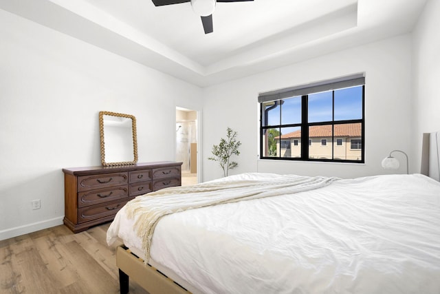 bedroom featuring baseboards, light wood finished floors, ceiling fan, a raised ceiling, and connected bathroom