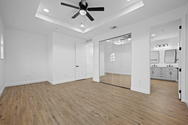 unfurnished bedroom featuring light wood-type flooring, ceiling fan, ensuite bathroom, and a tray ceiling