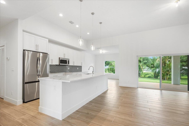 kitchen with white cabinetry, appliances with stainless steel finishes, a kitchen island with sink, decorative light fixtures, and light stone countertops