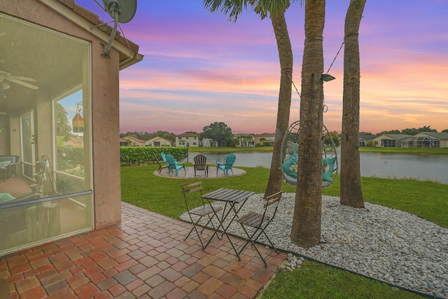 patio terrace at dusk featuring a fire pit, a lawn, ceiling fan, and a water view