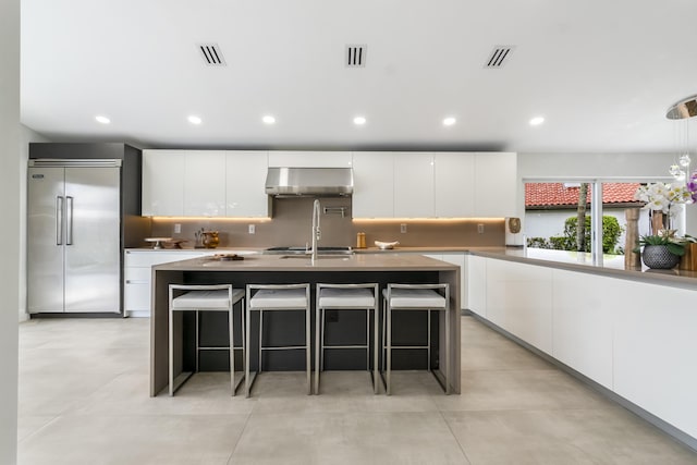 kitchen with a kitchen island with sink, backsplash, white cabinetry, a kitchen bar, and built in fridge