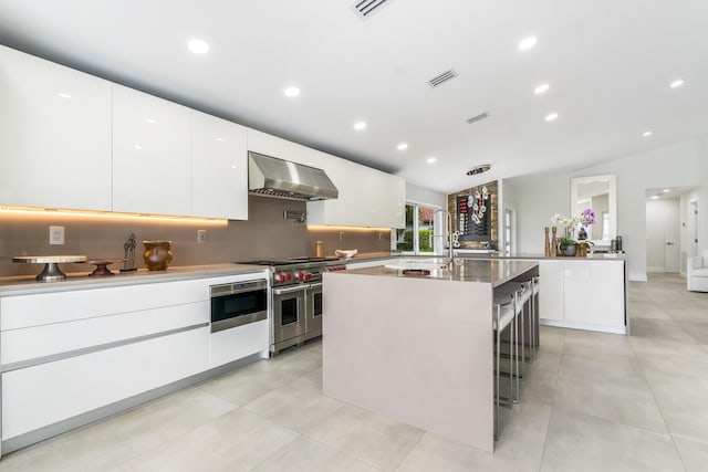 kitchen with wall chimney exhaust hood, sink, vaulted ceiling, stainless steel appliances, and a kitchen island with sink