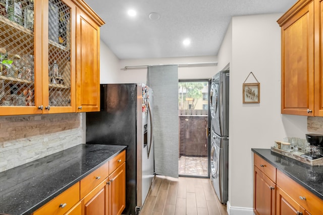 kitchen with stainless steel fridge, dark stone counters, stacked washer and clothes dryer, light hardwood / wood-style floors, and decorative backsplash