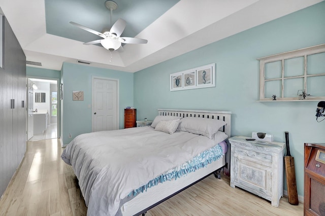 bedroom featuring ceiling fan, connected bathroom, light hardwood / wood-style floors, and a tray ceiling