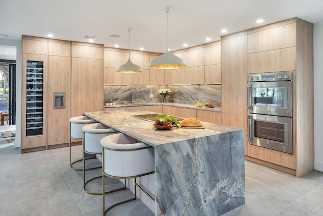 kitchen featuring light stone counters, light brown cabinets, double oven, and hanging light fixtures