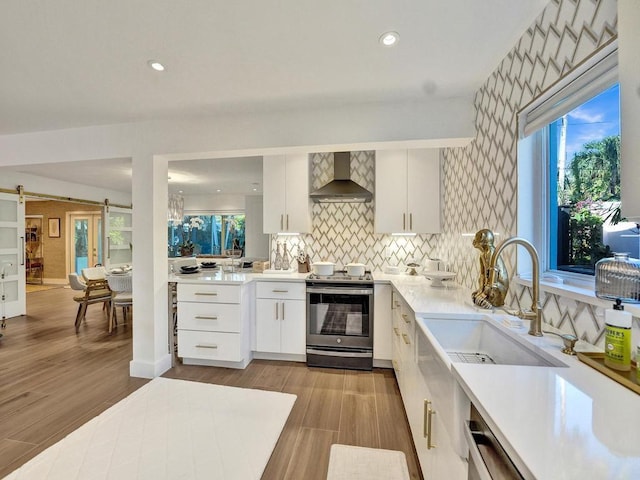 kitchen with a barn door, wall chimney range hood, white cabinetry, plenty of natural light, and stainless steel electric stove