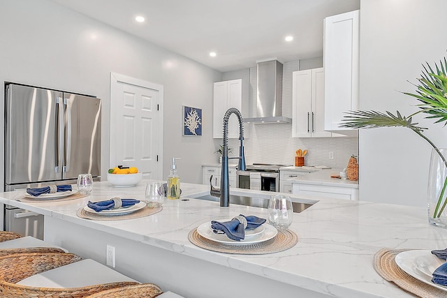 kitchen with light stone counters, wall chimney range hood, white cabinetry, and stainless steel fridge