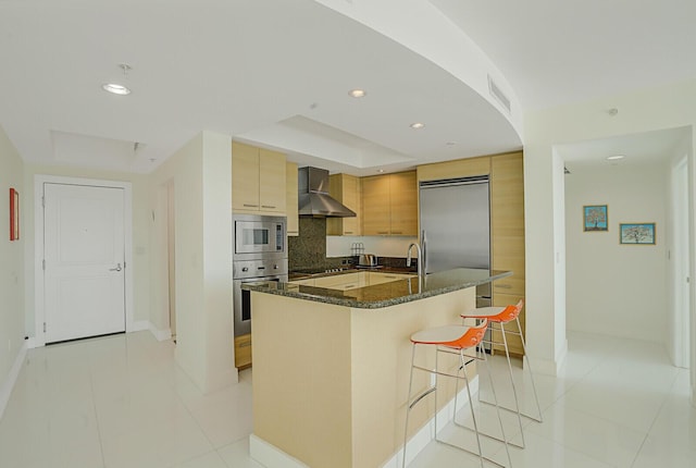 kitchen with wall chimney range hood, a breakfast bar area, dark stone countertops, built in appliances, and an island with sink