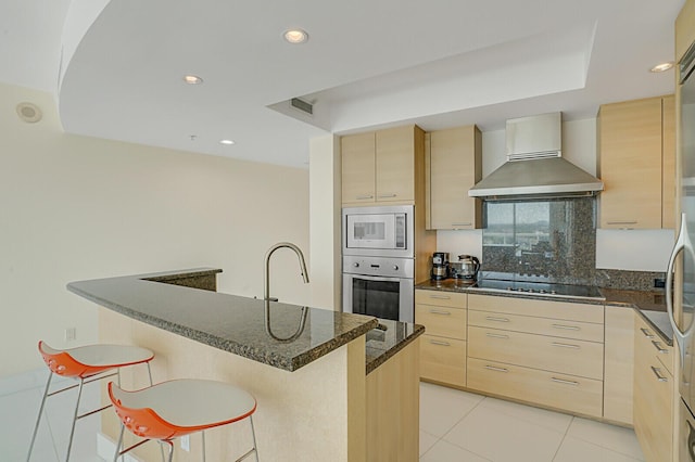 kitchen featuring light brown cabinetry, dark stone countertops, a kitchen breakfast bar, stainless steel appliances, and wall chimney exhaust hood