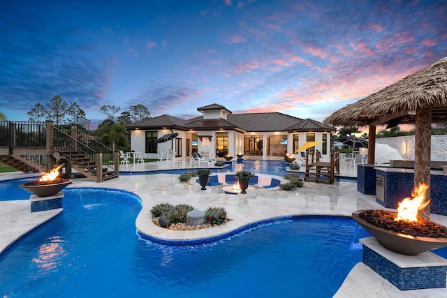 pool at dusk featuring an outdoor kitchen, a patio area, pool water feature, and an outdoor fire pit