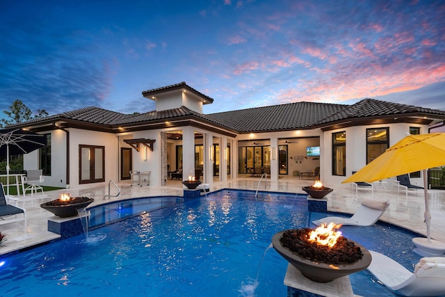 pool at dusk featuring a fire pit, pool water feature, and ceiling fan