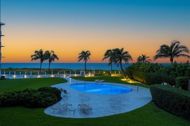 pool at dusk featuring a water view, a yard, and a patio area