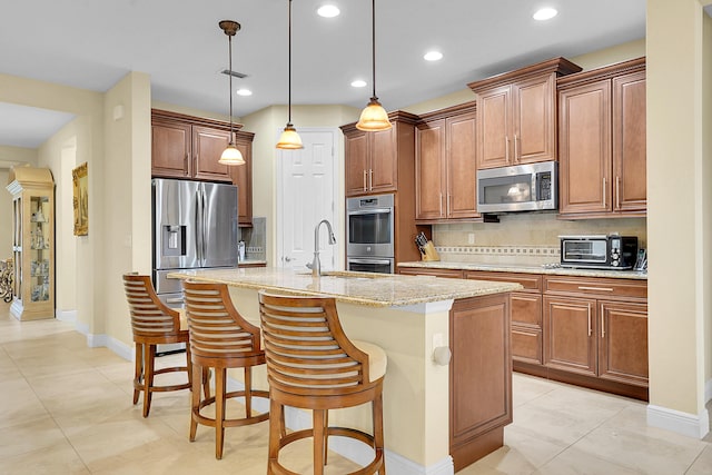 kitchen featuring appliances with stainless steel finishes, tasteful backsplash, decorative light fixtures, an island with sink, and light stone counters