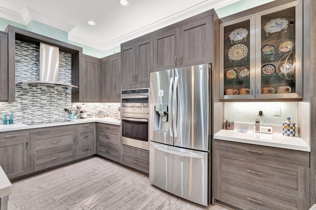 kitchen featuring crown molding, tasteful backsplash, light wood-type flooring, stainless steel appliances, and wall chimney range hood