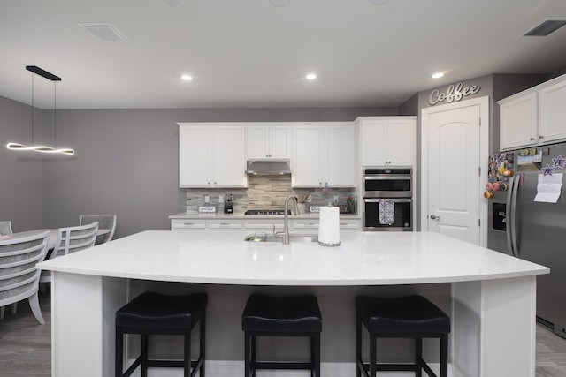 kitchen featuring appliances with stainless steel finishes, a kitchen island with sink, white cabinetry, light stone counters, and decorative backsplash