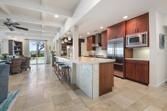 kitchen featuring wall chimney exhaust hood, a breakfast bar, an island with sink, beamed ceiling, and built in appliances