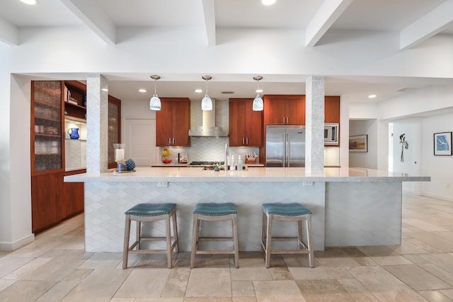 kitchen featuring decorative light fixtures, wall chimney range hood, built in appliances, light stone counters, and a breakfast bar area