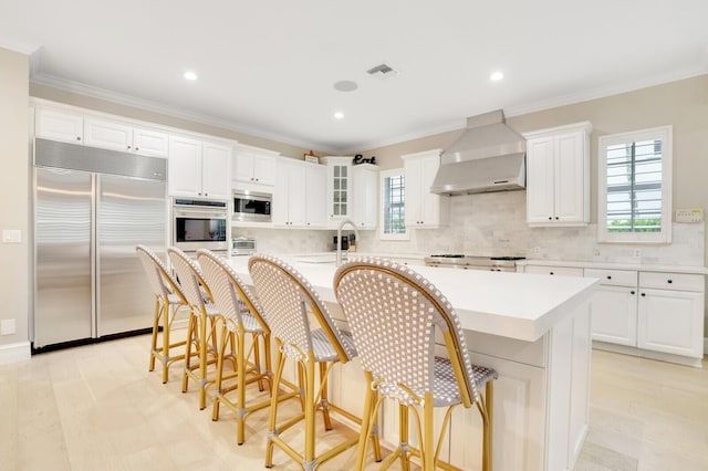 kitchen with built in appliances, a center island, white cabinetry, a kitchen breakfast bar, and wall chimney range hood