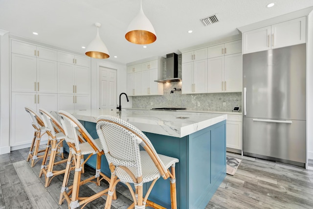 kitchen featuring stainless steel refrigerator, white cabinetry, decorative light fixtures, and wall chimney range hood