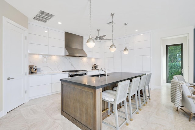 kitchen featuring wall chimney exhaust hood, sink, white cabinetry, pendant lighting, and a kitchen island with sink