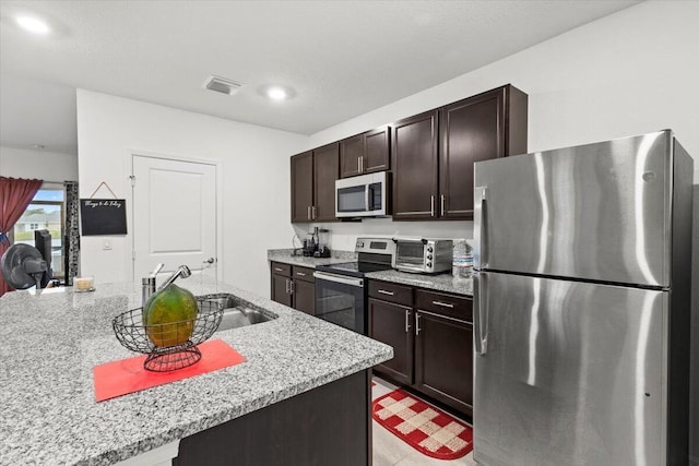 kitchen featuring a toaster, stainless steel appliances, a sink, dark brown cabinetry, and light stone countertops