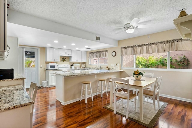 kitchen with white cabinets, a kitchen bar, kitchen peninsula, light stone countertops, and white appliances