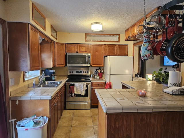 kitchen featuring tile countertops, a textured ceiling, stainless steel appliances, a sink, and brown cabinets