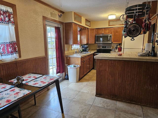 kitchen featuring tile countertops, a textured ceiling, a wainscoted wall, stainless steel appliances, and brown cabinets