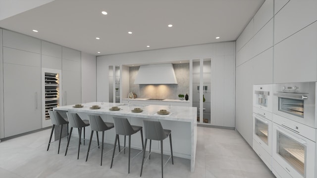 kitchen featuring white cabinets, a kitchen bar, a kitchen island with sink, light stone counters, and custom range hood