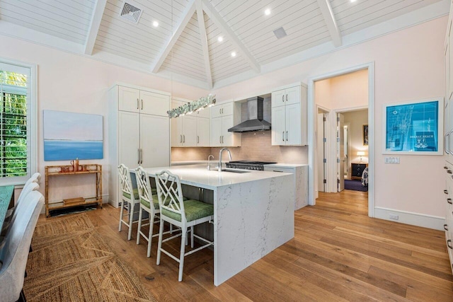 kitchen with wall chimney exhaust hood, tasteful backsplash, a center island with sink, beam ceiling, and white cabinets