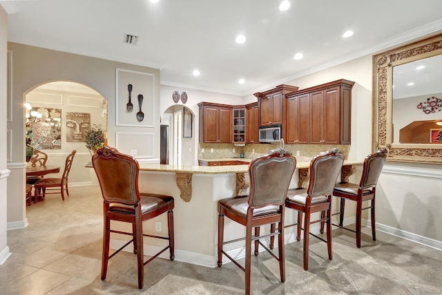 kitchen with a breakfast bar area, backsplash, light stone countertops, ornamental molding, and kitchen peninsula