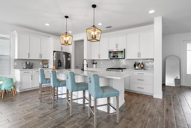 kitchen with appliances with stainless steel finishes, an island with sink, sink, white cabinets, and hanging light fixtures