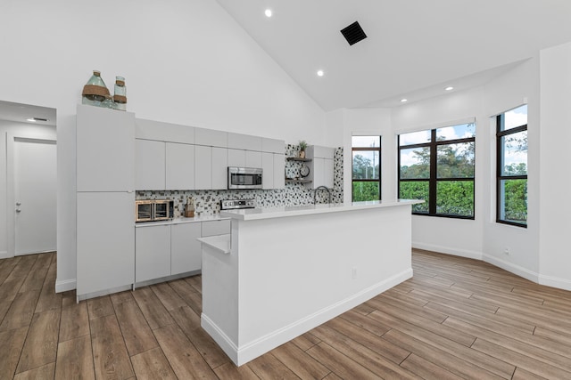 kitchen featuring high vaulted ceiling, white cabinetry, backsplash, stainless steel appliances, and a center island with sink